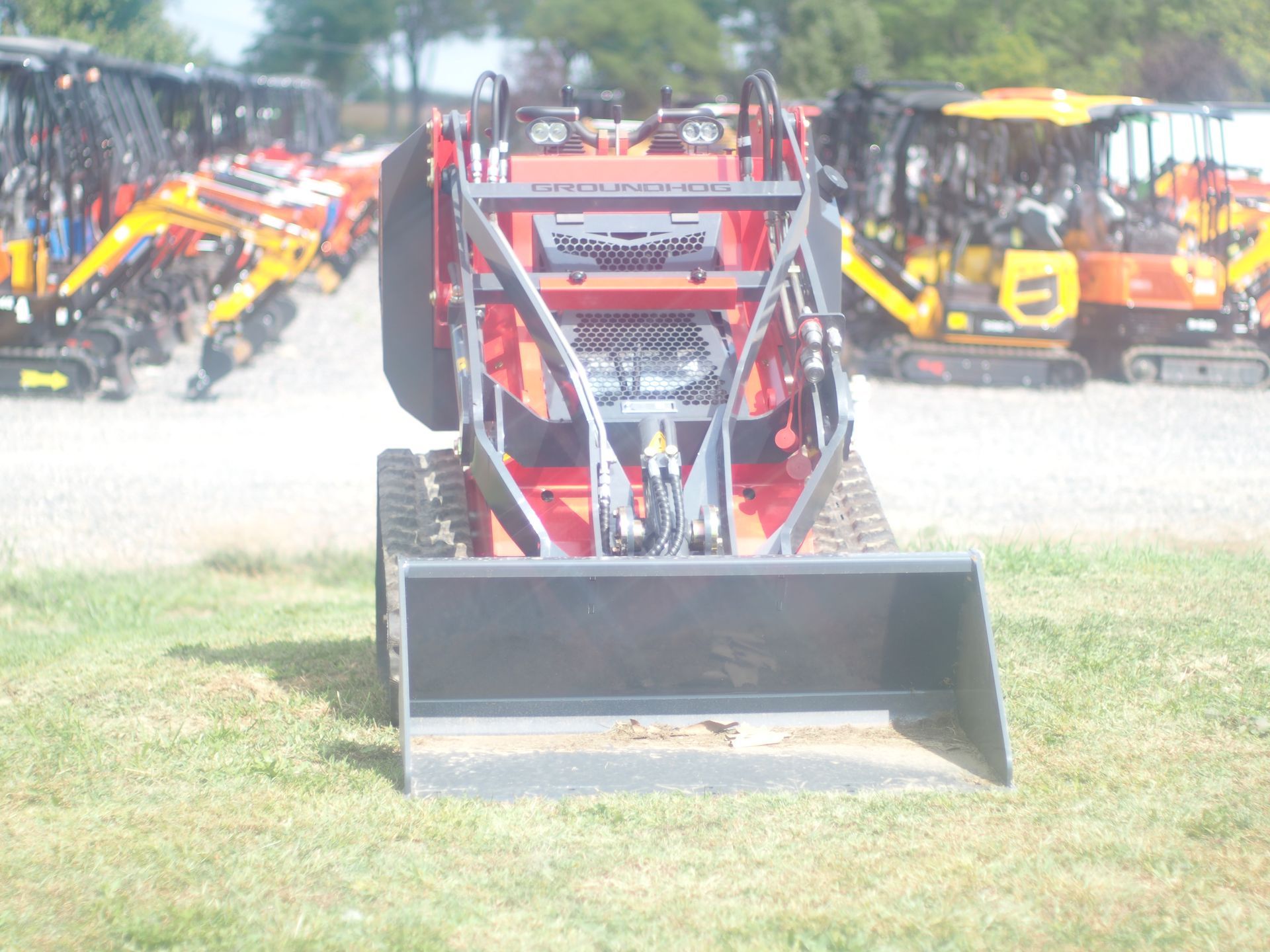 Red skid steer loader with a black bucket on grass, other equipment in the background.