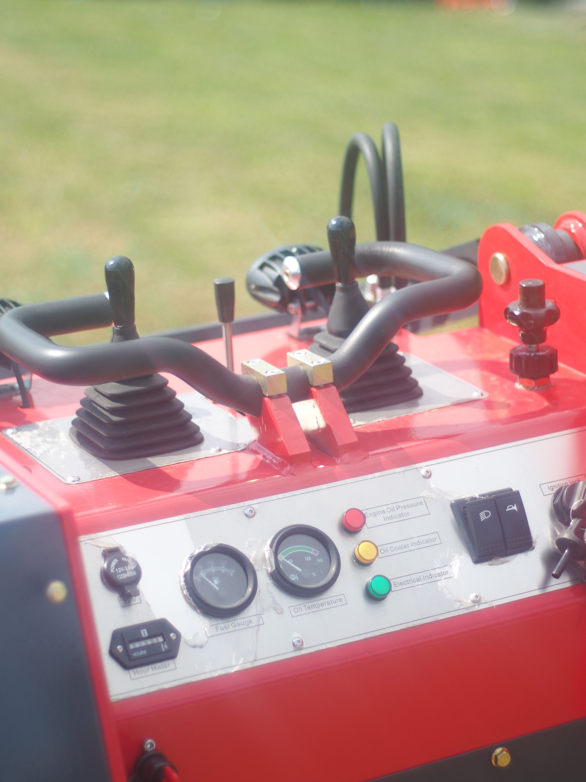 Control panel of a red and silver machine with gauges, levers, and buttons; outdoors on grass.