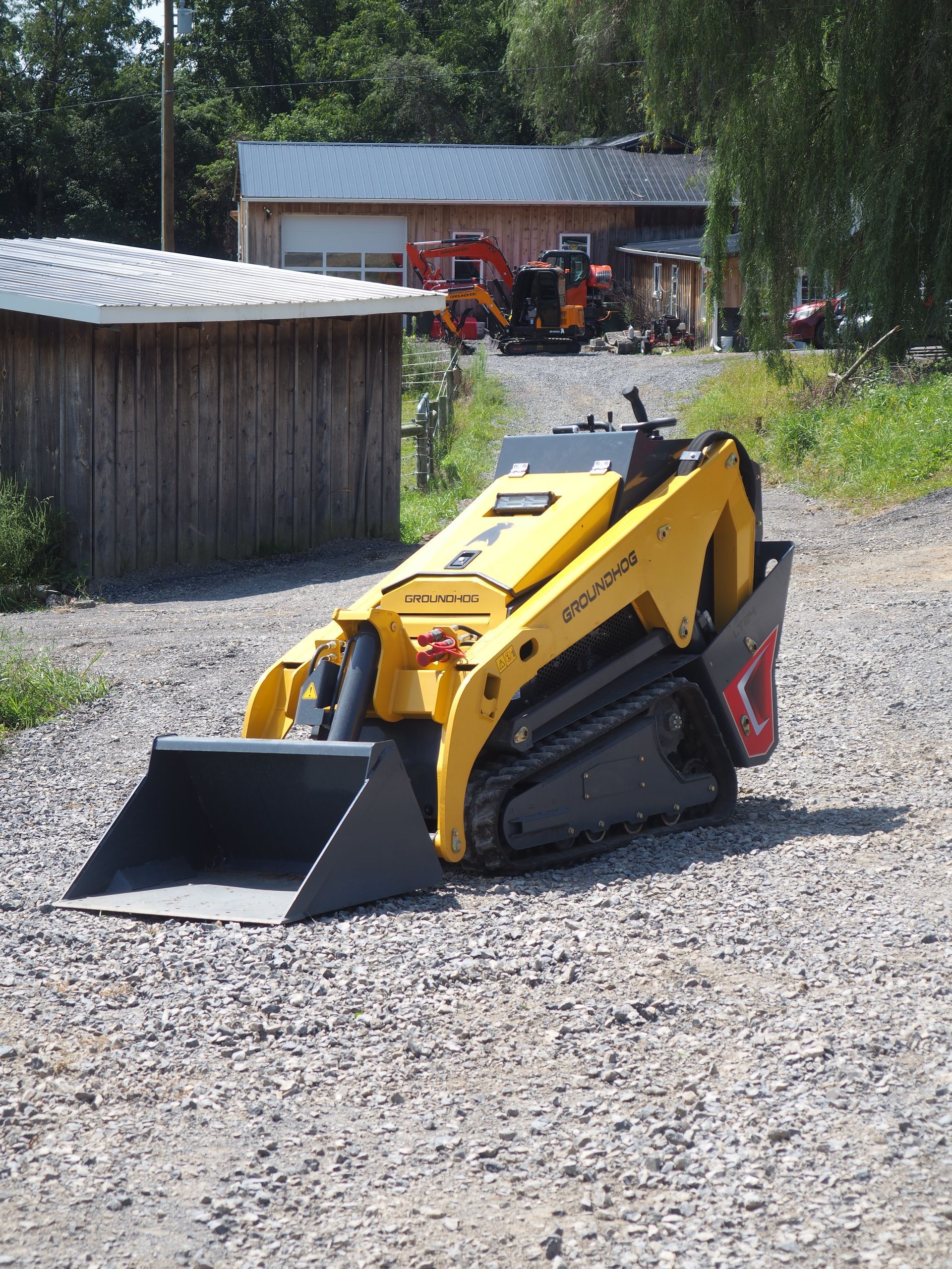 Yellow compact track loader on gravel driveway, with wooden shed and building in background.