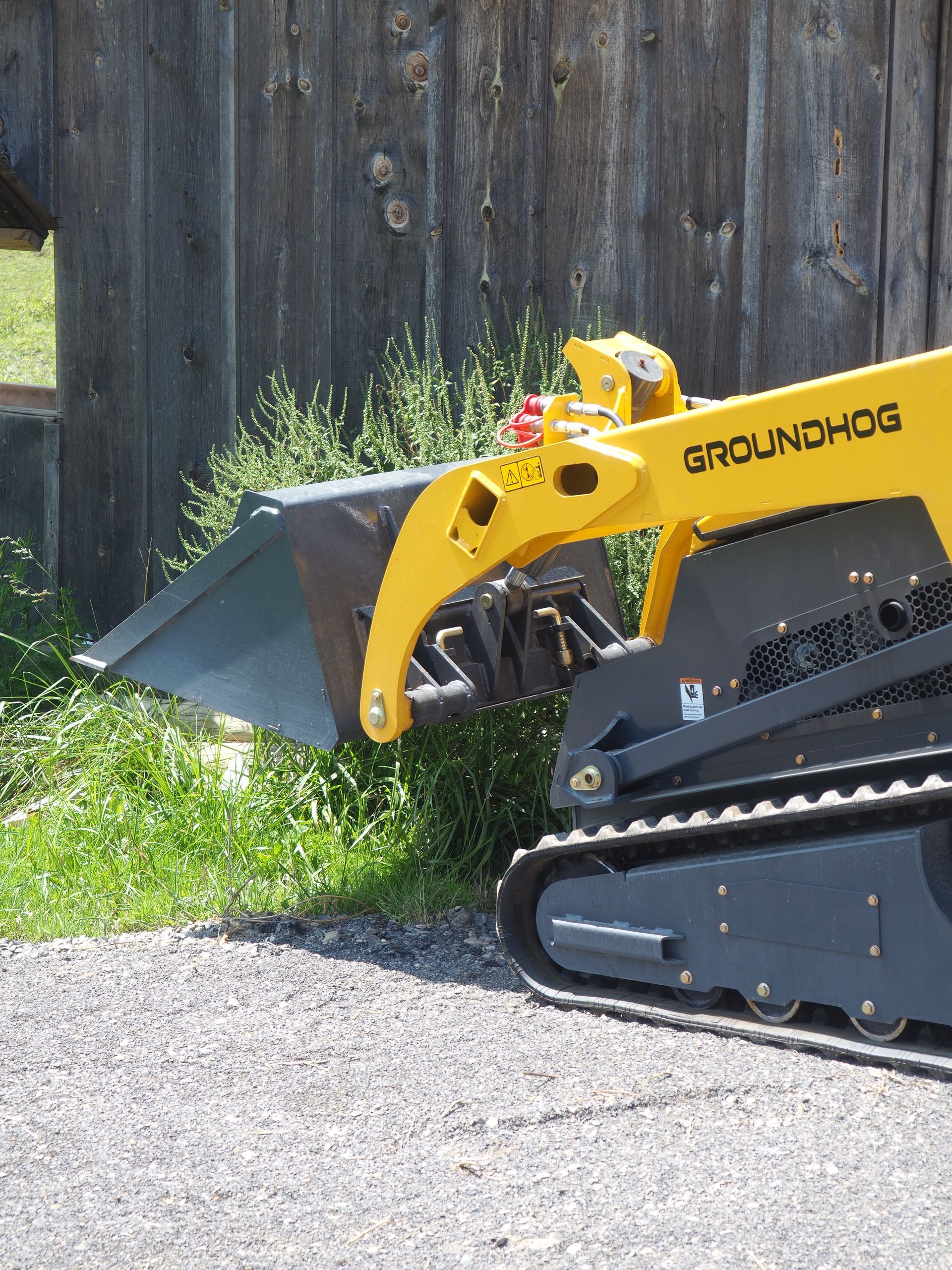 Yellow Groundhog mini skid steer loader with bucket, parked on gravel in front of a wooden fence.