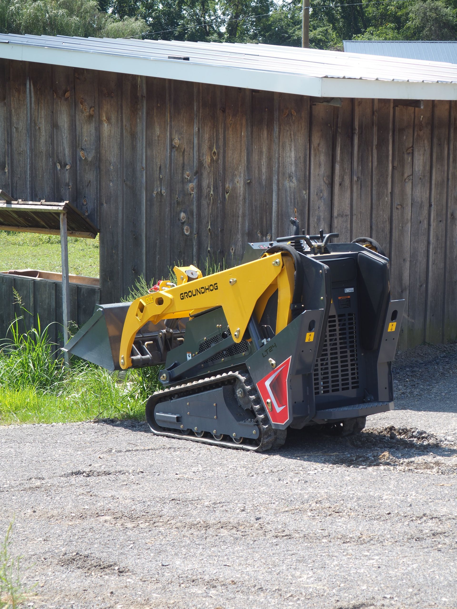 Yellow and black compact track loader parked on gravel in front of a wooden building.