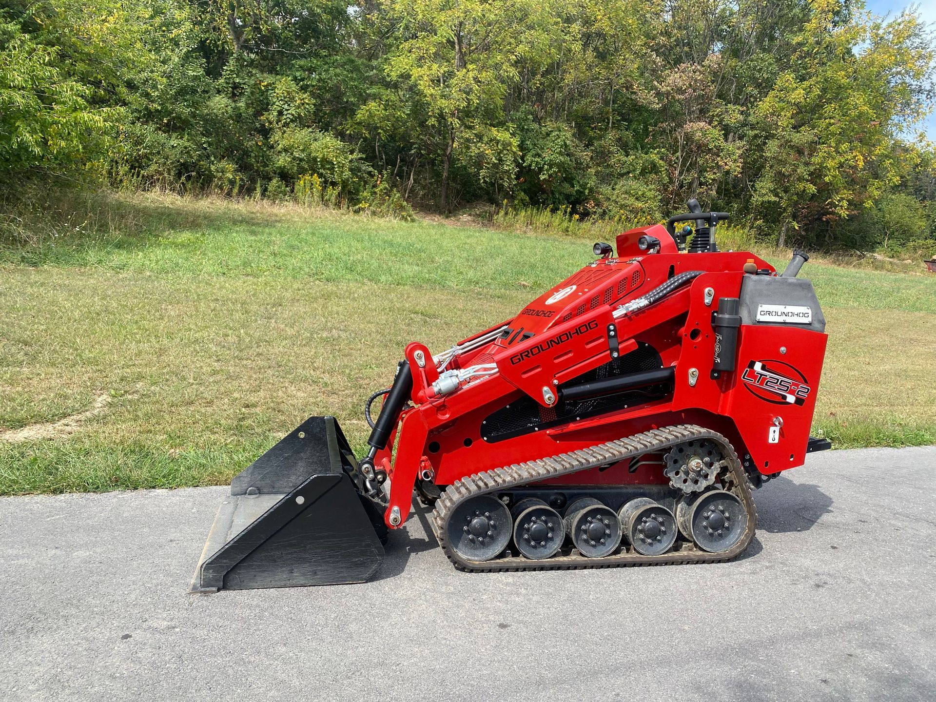 Red tracked mini skid steer with a bucket on pavement near grass and trees.