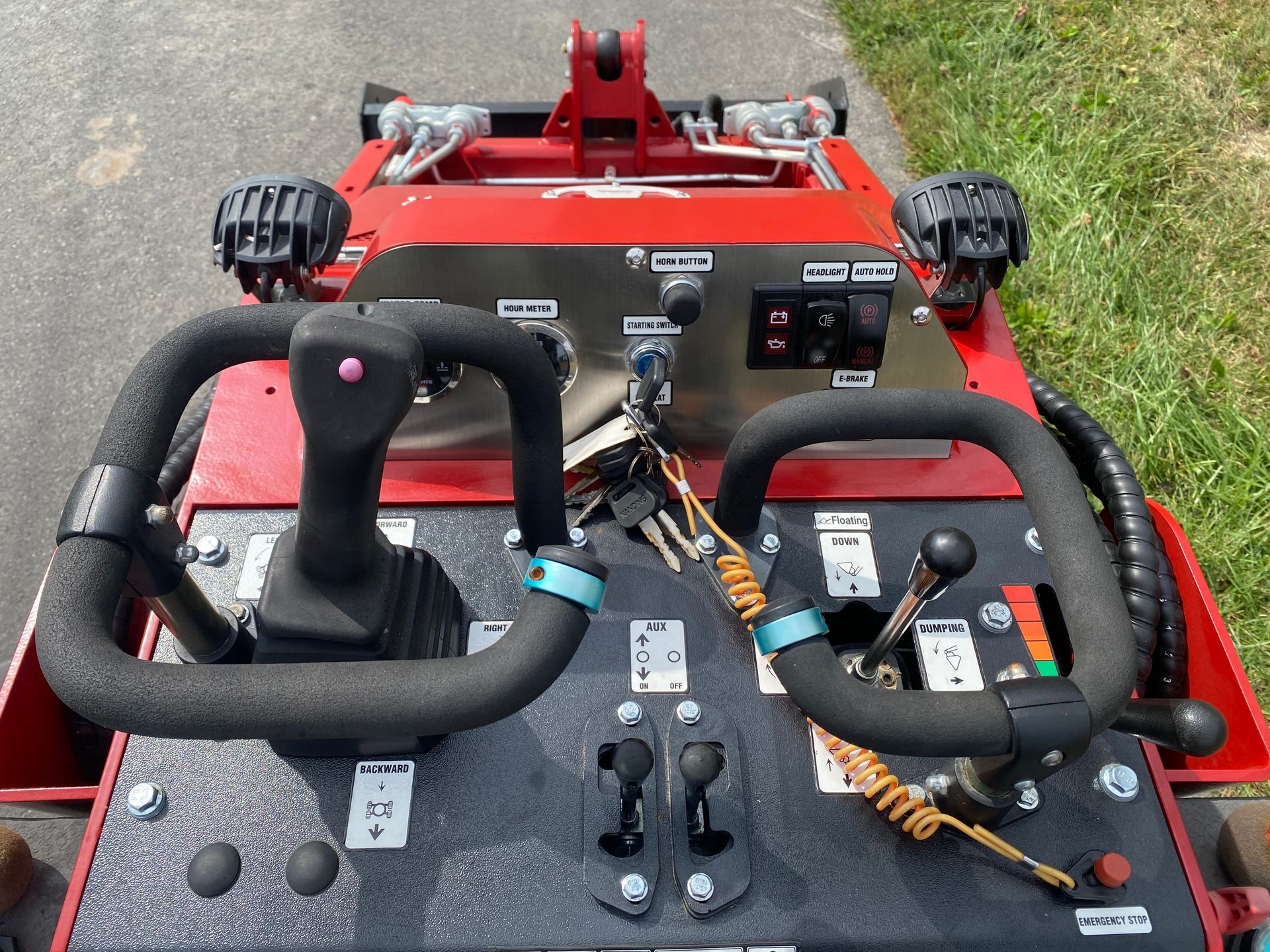 Red and black control panel of a machine, with joysticks, handles, and switches, on a paved surface.
