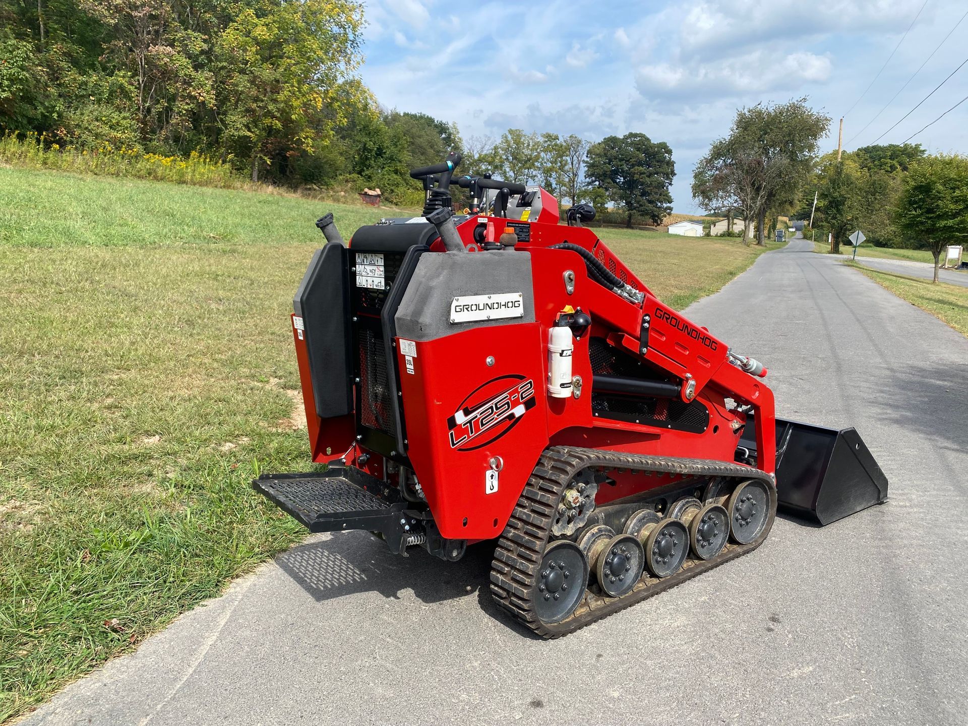 Red mini skid steer loader on a paved road, with a field and trees in the background.