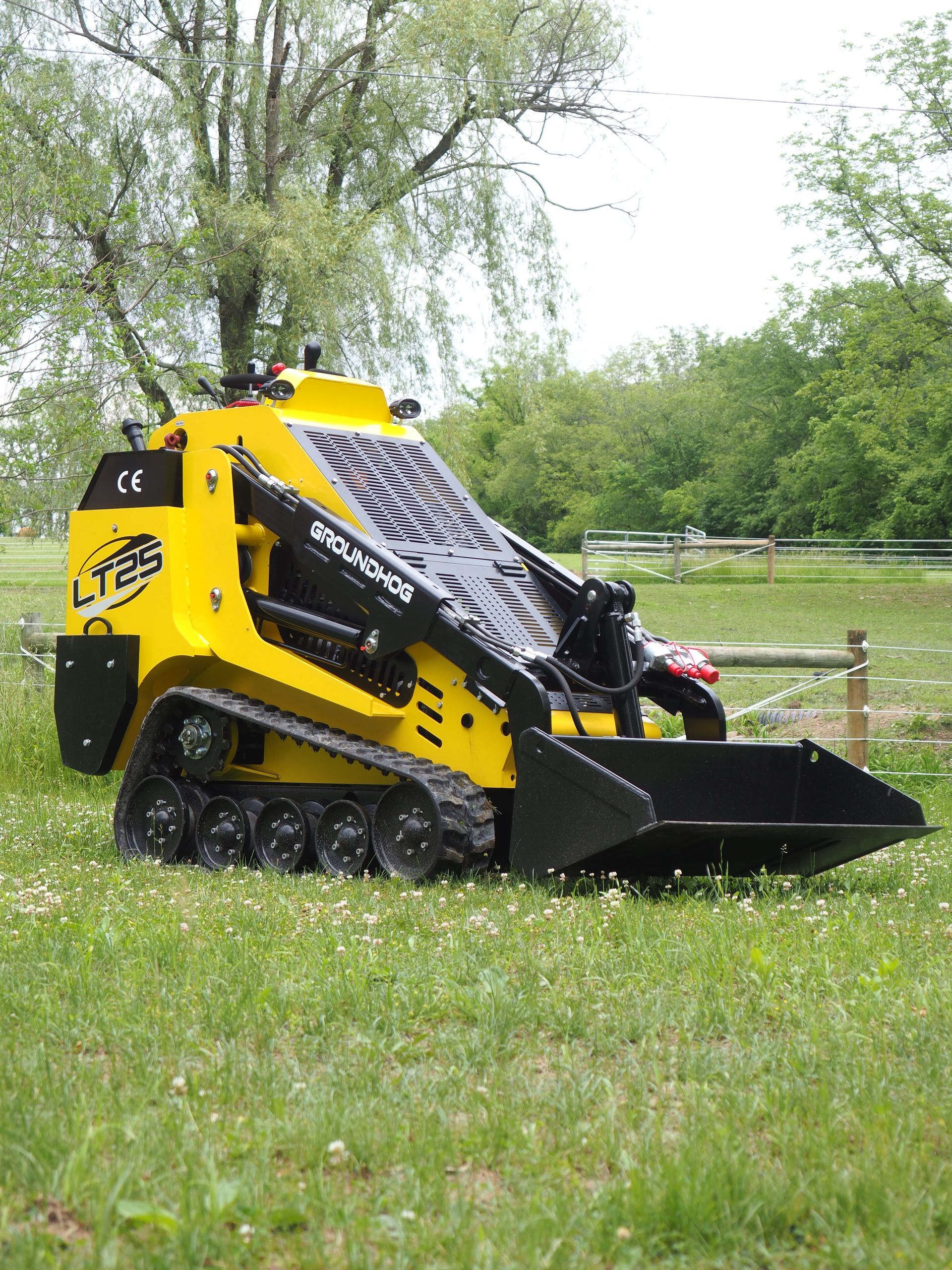 Yellow tracked skid steer loader with black bucket, on grass, near trees.