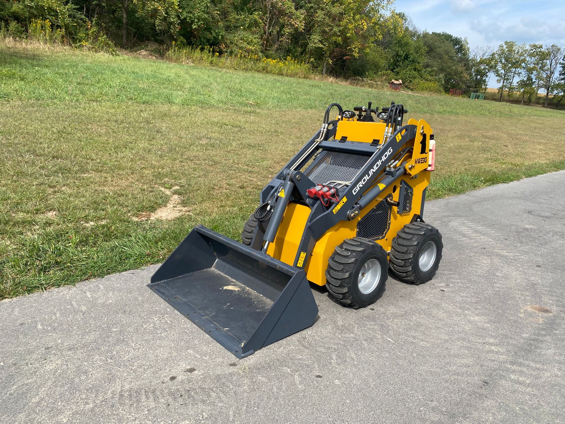 Yellow and black mini skid steer with a bucket on a paved surface, field in background.