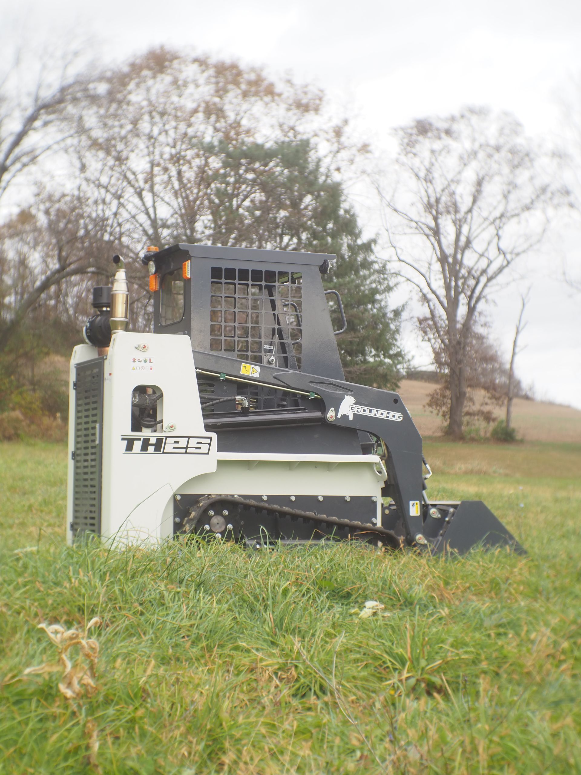 White and black tracked skid steer on a grassy field, in front of trees and an overcast sky.