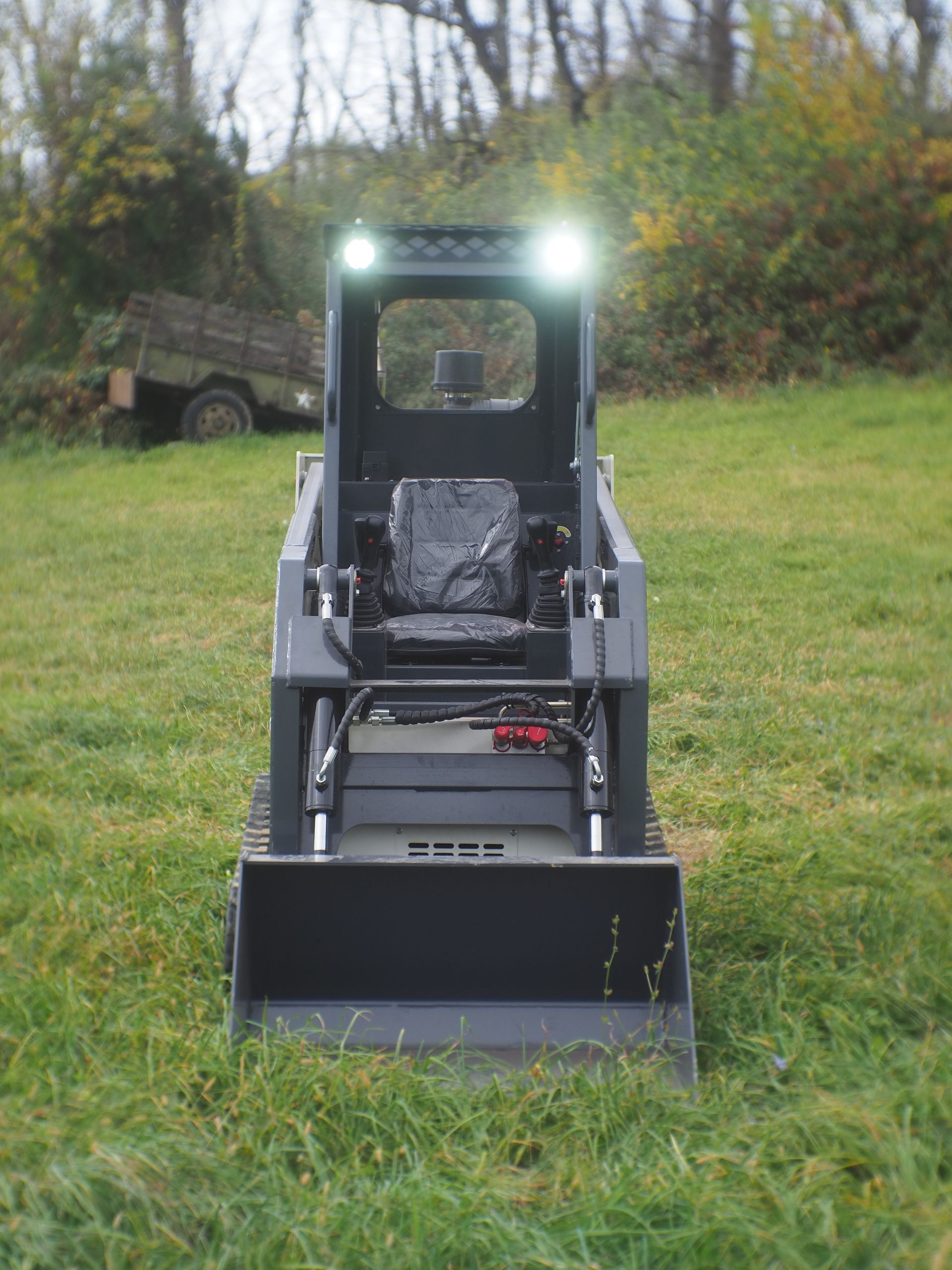 Gray skid steer loader on a grassy field with bright lights, a trailer, and trees in the background.