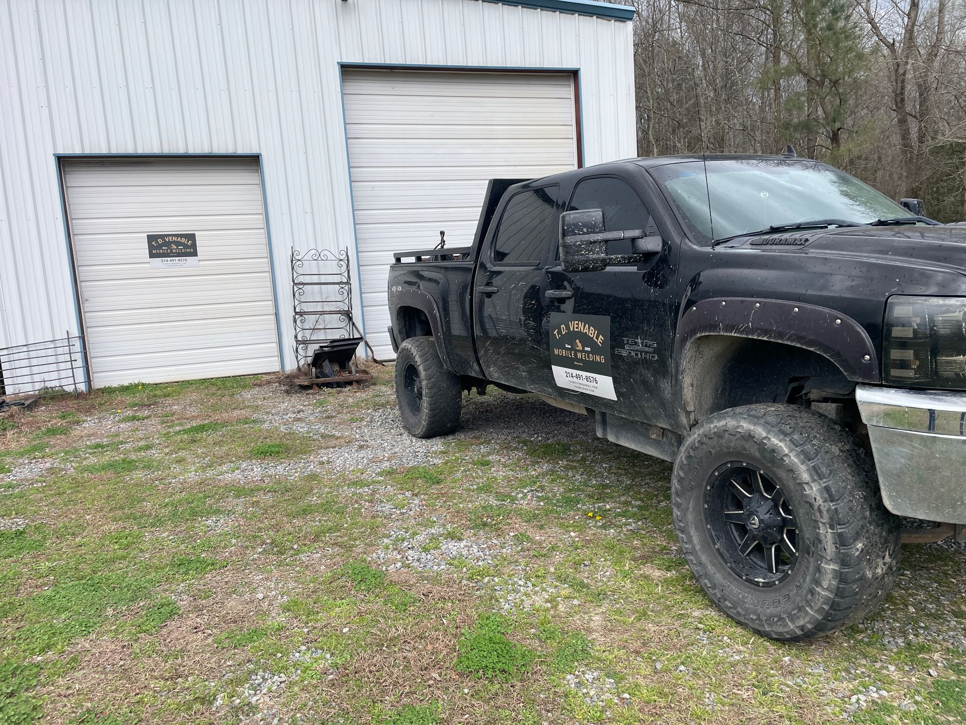 A black lifted pickup truck parked on gravel in front of a white metal industrial building with two garage doors.