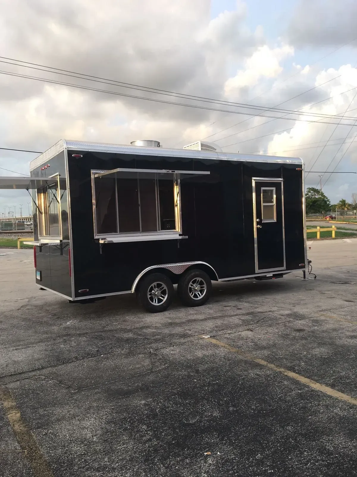 Black food trailer with serving window parked in a lot under cloudy sky.