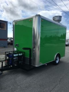 Green enclosed cargo trailer with a rear-mounted generator parked on pavement under a cloudy sky