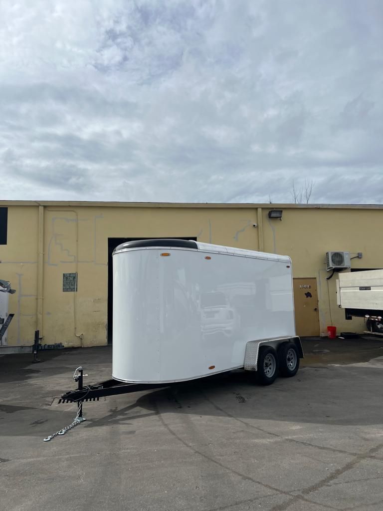 White enclosed cargo trailer parked in an empty lot beside a beige building under cloudy sky