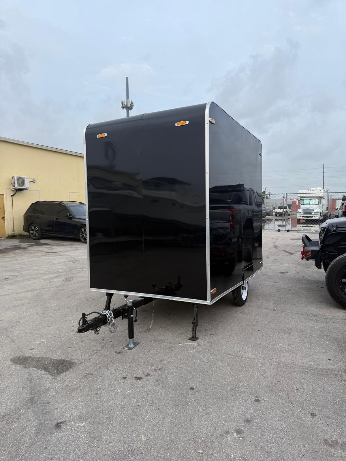 Black enclosed trailer with hitch, parked in a lot beside vehicles and a building.