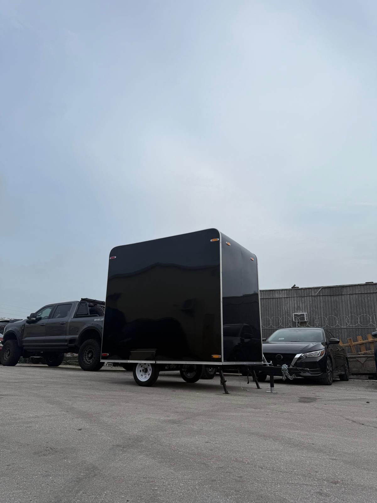 Black enclosed cargo trailer parked in a lot, with a pickup truck and car in the background.