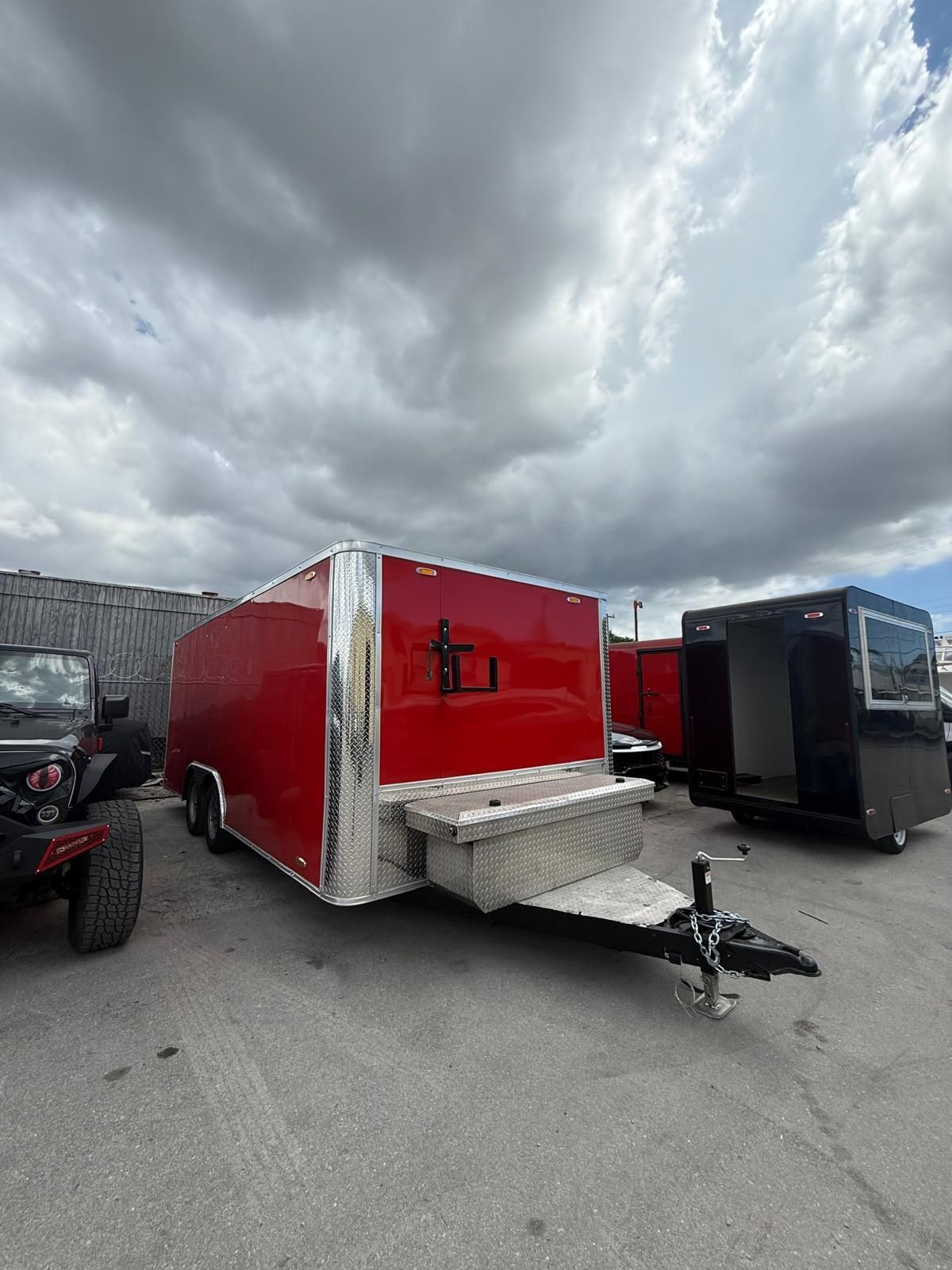 Red enclosed utility trailer with open rear ramp parked on a lot under cloudy skies.