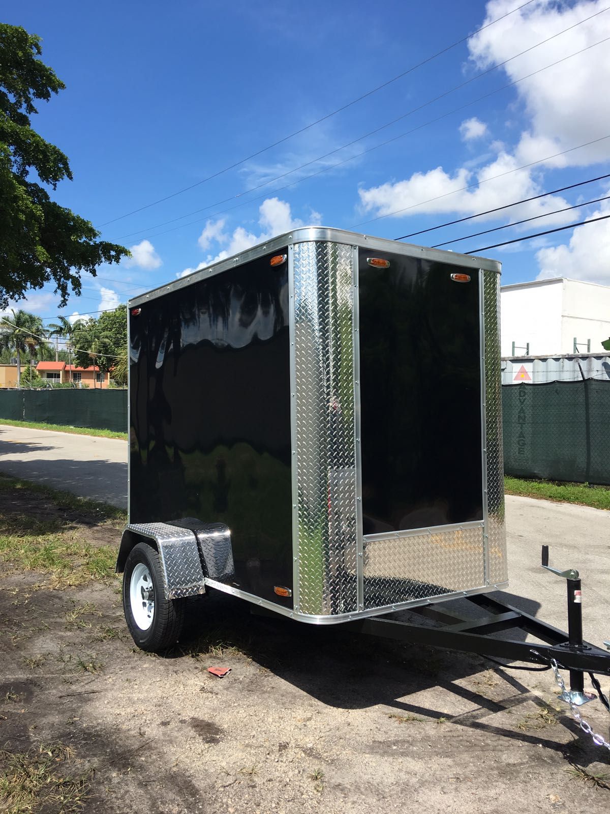 Black enclosed cargo trailer with silver trim parked on a gravel lot under a blue sky.