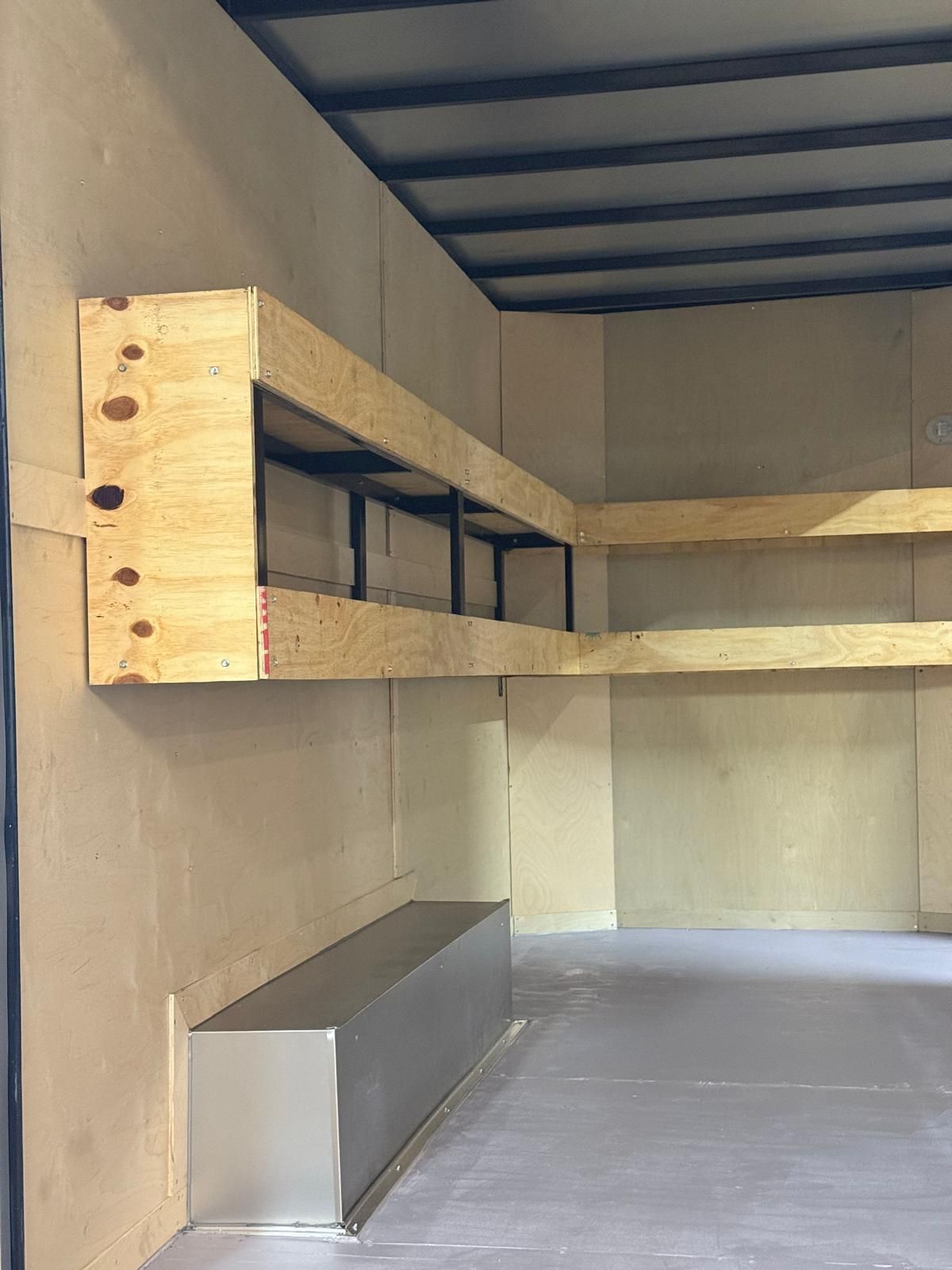 Interior of a small storage compartment with two wooden shelves, gray floor, and unfinished walls.