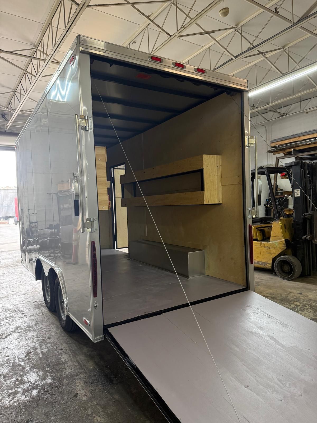 Open cargo trailer with ramp door down inside a warehouse, showing empty interior and shelves.