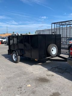 Black utility trailer with spare tire parked in an outdoor lot under a blue sky.
