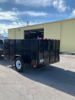 Black utility trailer parked outside a beige warehouse on a sunny day.