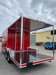 Red enclosed trailer with open rear ramp door, parked in an industrial lot under cloudy sky.
