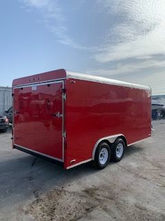 Red enclosed cargo trailer with tandem axles parked on pavement under a cloudy sky.