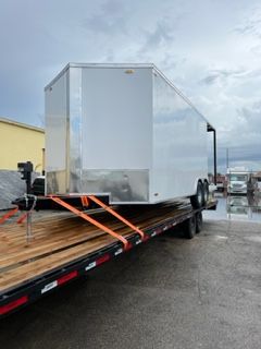 White cargo trailer strapped to a flatbed truck in a wet parking lot under cloudy skies