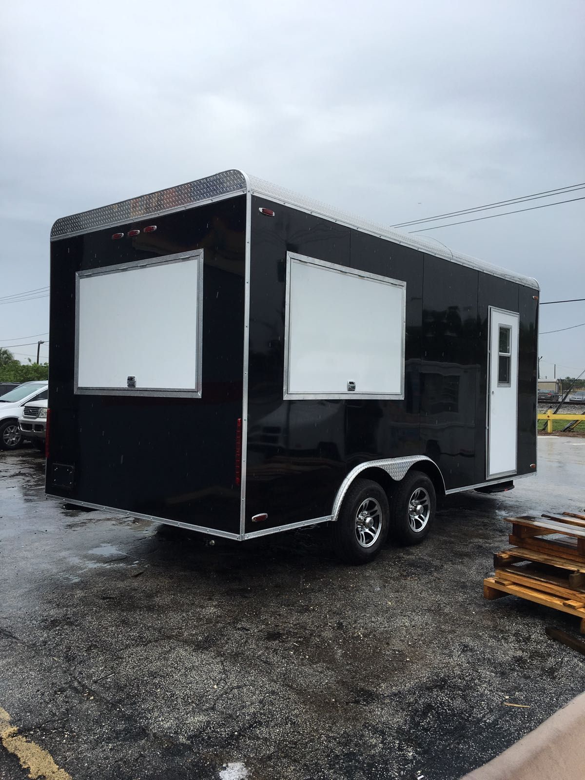 Black mobile trailer with white service windows, parked on wet gravel under cloudy sky.
