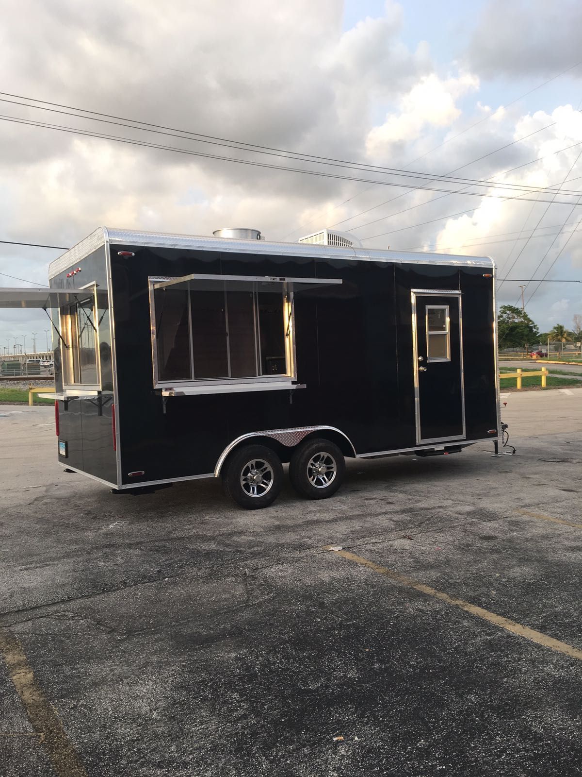 Black food trailer with serving window and door parked on a paved lot under cloudy sky