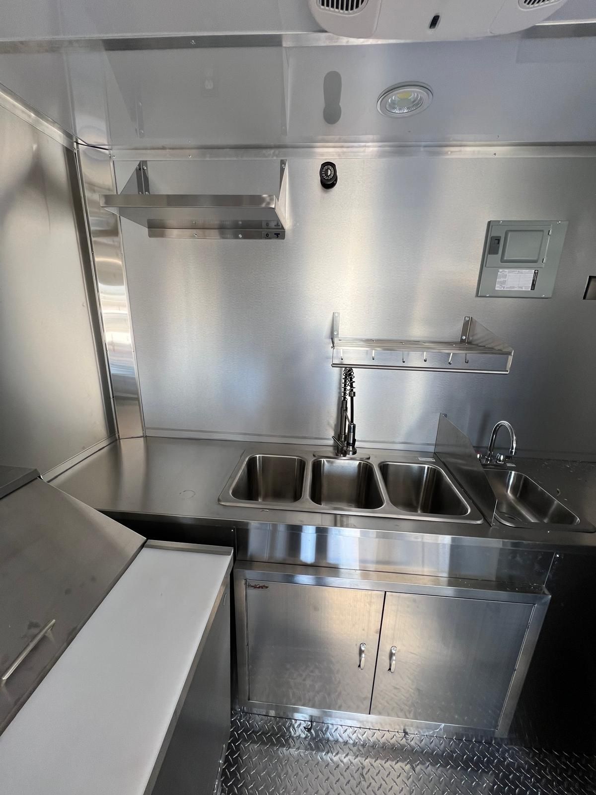 Compact stainless-steel kitchen sink area with double basins, faucet, and wall-mounted dispenser.