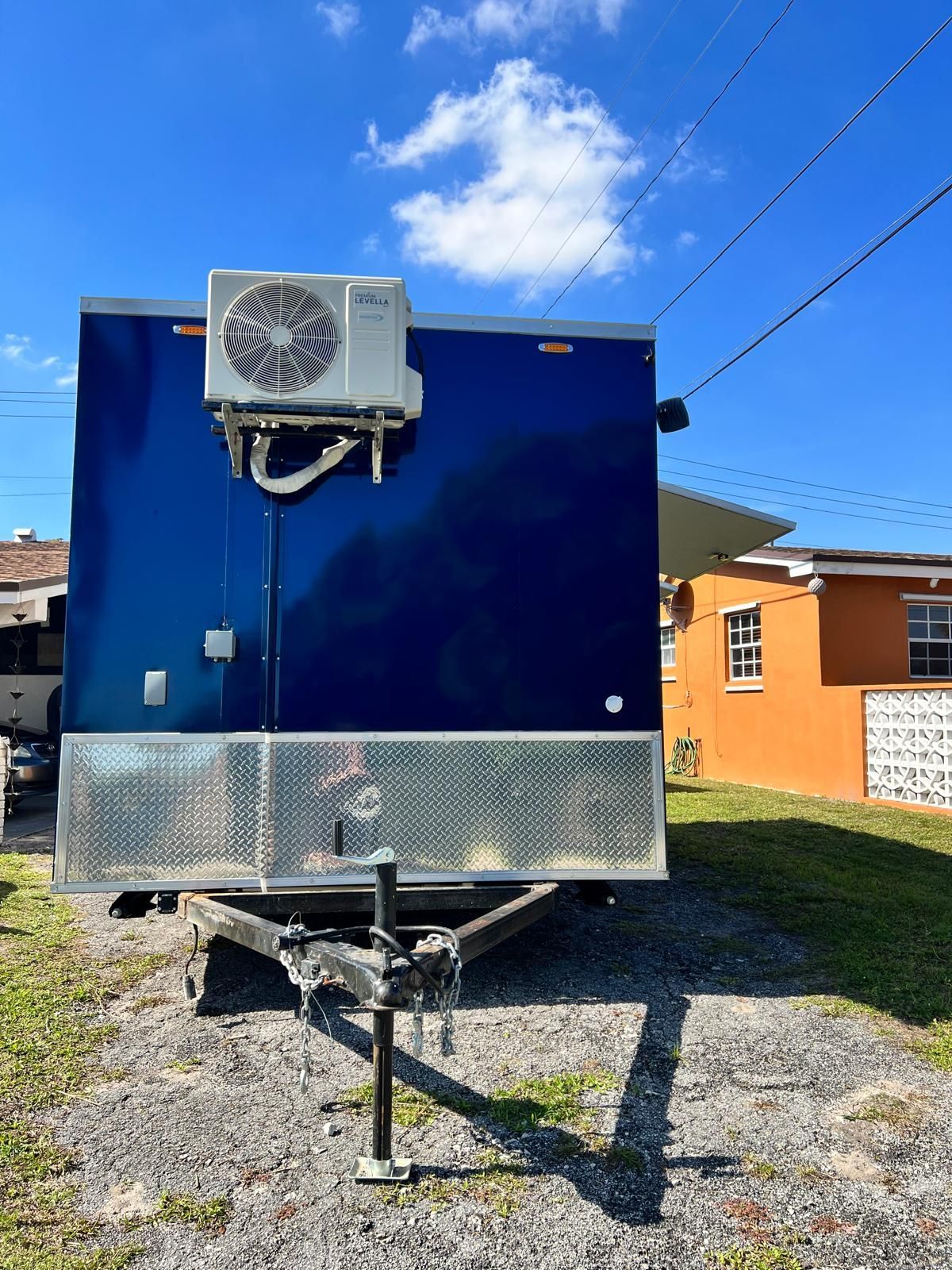 Blue trailer with front-mounted air conditioner parked on a gravel lot under a blue sky