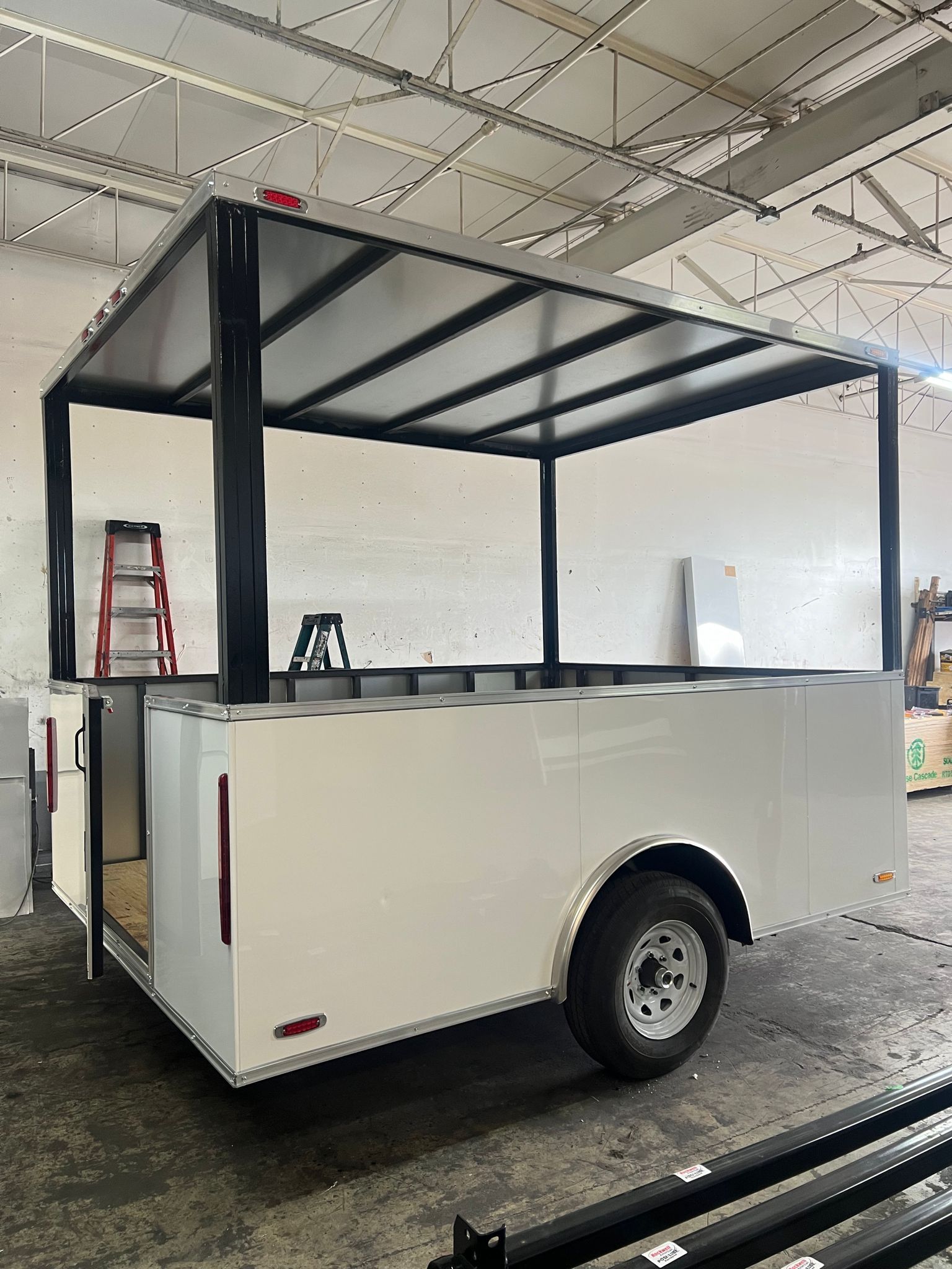 White utility trailer with black metal roof frame in a warehouse workshop.