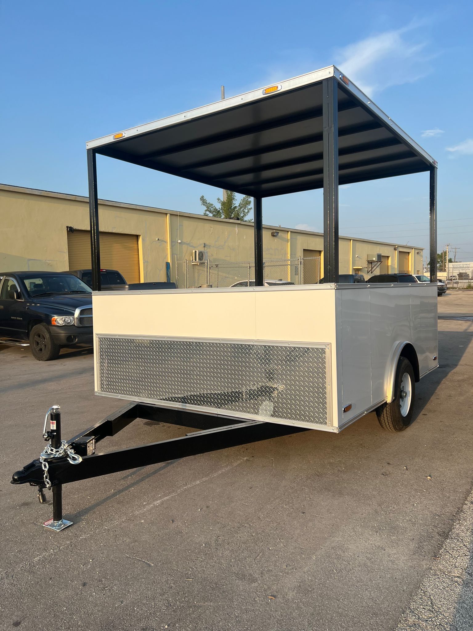 White utility trailer with canopy roof, front hitch, and diamond-plate paneling parked outdoors in a lot.