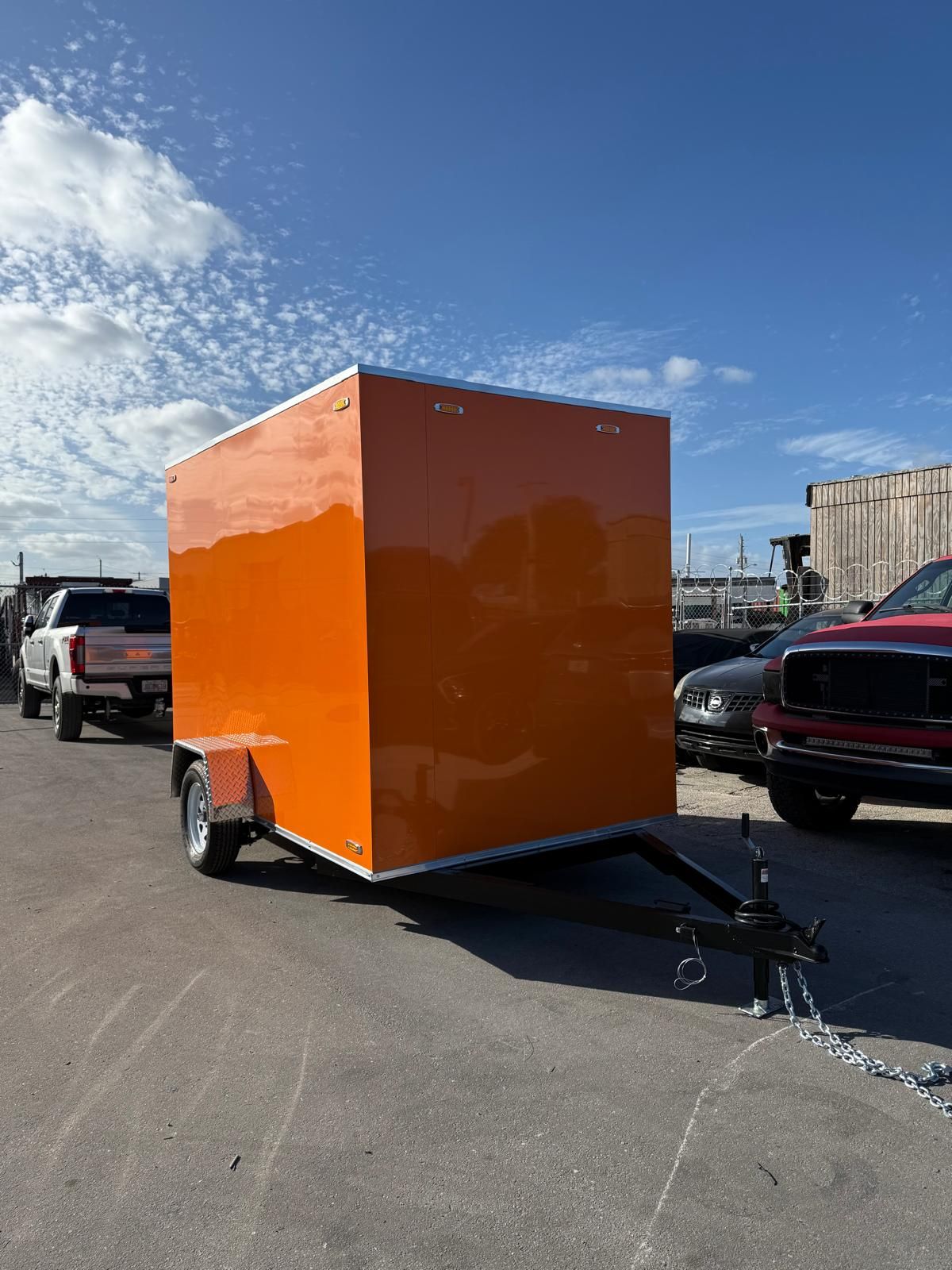 Bright orange enclosed trailer parked in a lot under a blue sky, hitched to a vehicle.