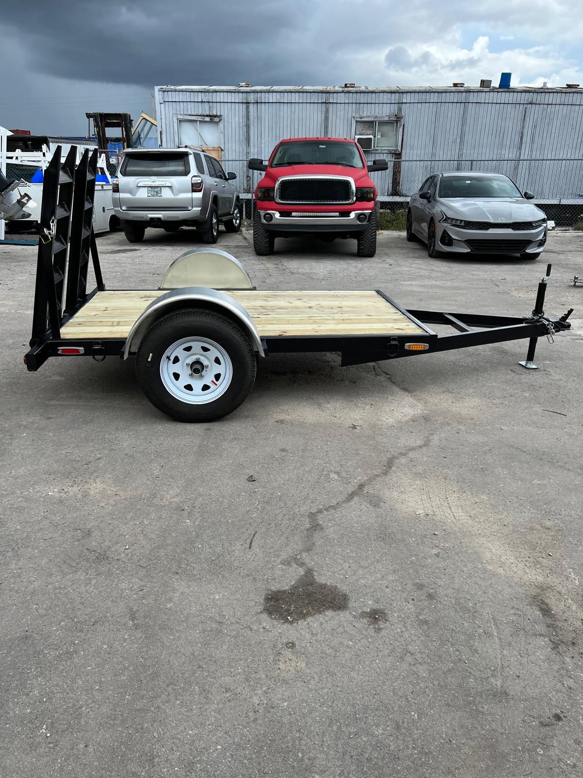Empty utility trailer with wooden deck and black frame parked in an auto lot, cars in background