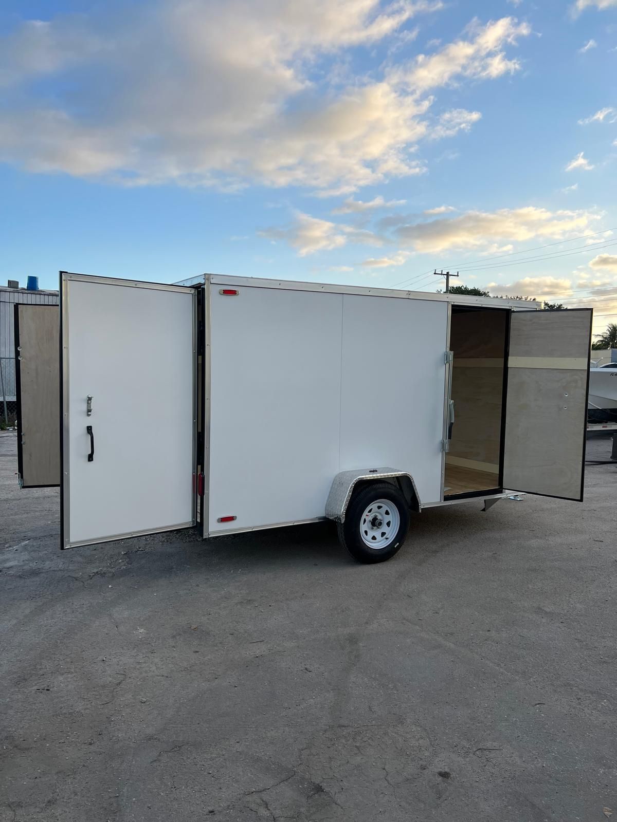White enclosed trailer with side doors open, parked on pavement under a partly cloudy sky.