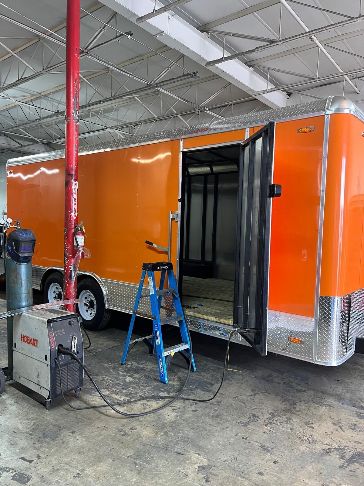 Orange enclosed trailer in a workshop, with an open side door, blue step ladder, and equipment nearby.