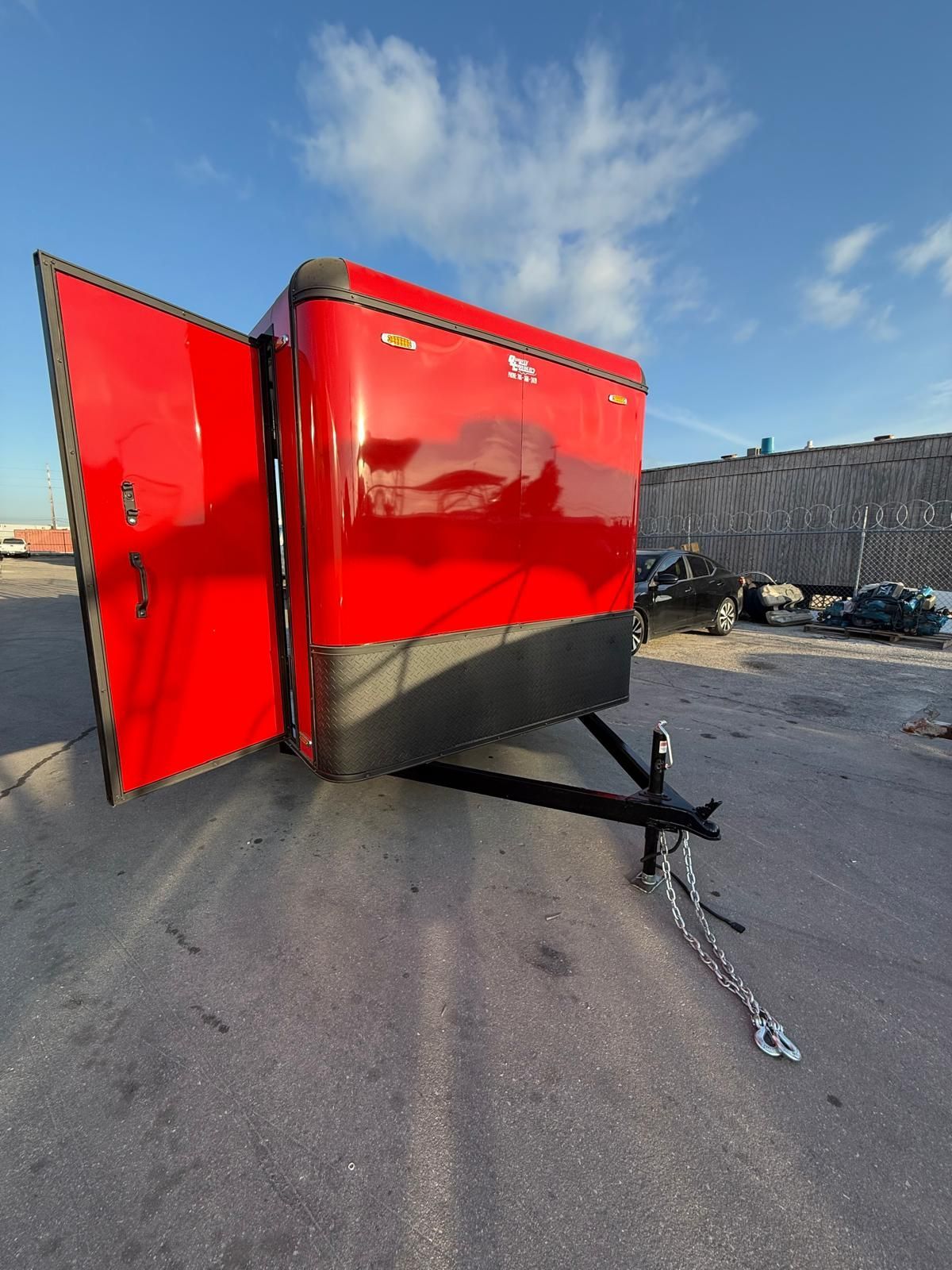 Red enclosed trailer with open door, parked on pavement with a chain and hitch visible under a blue sky
