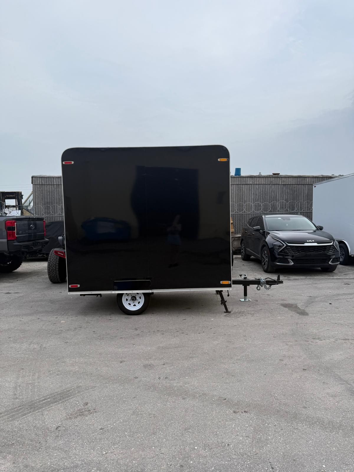 Black enclosed trailer parked in a lot, with a car and truck nearby under a cloudy sky.