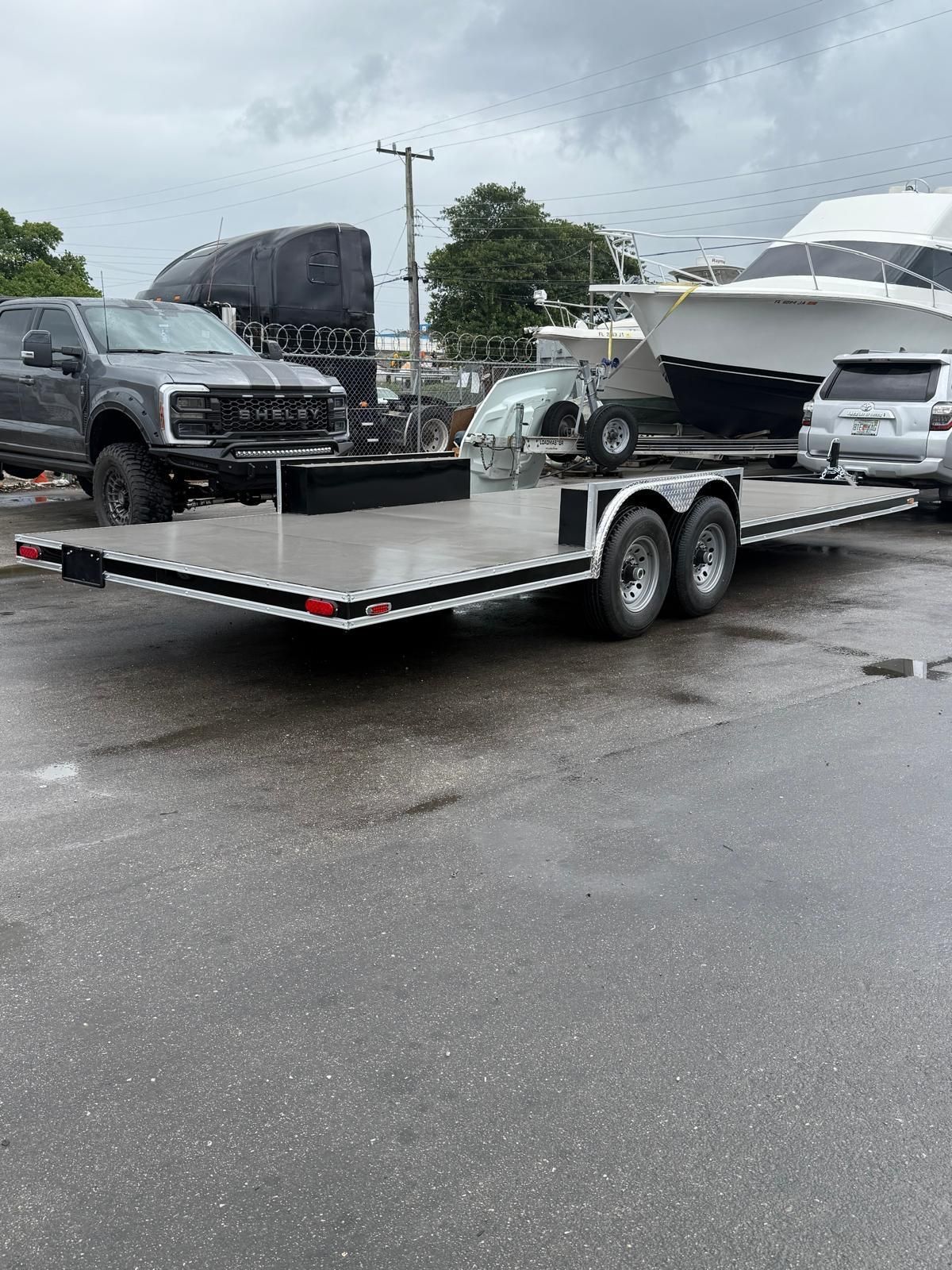 Silver dual-axle trailer with a black truck and white boat parked on a wet lot under cloudy skies