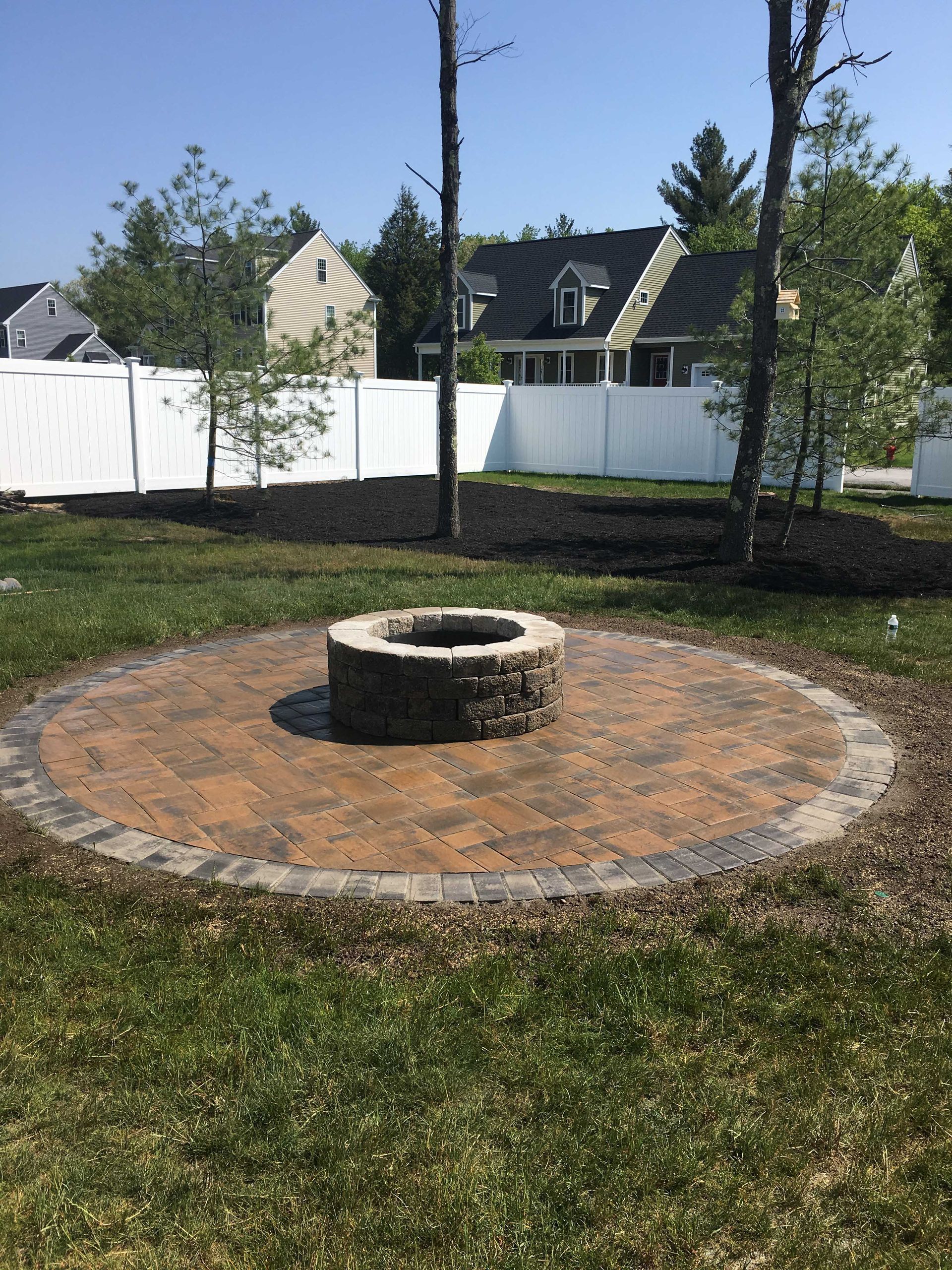 A circular stone fire pit sits on a brown paver patio in a backyard with a white fence, trees, and grass.