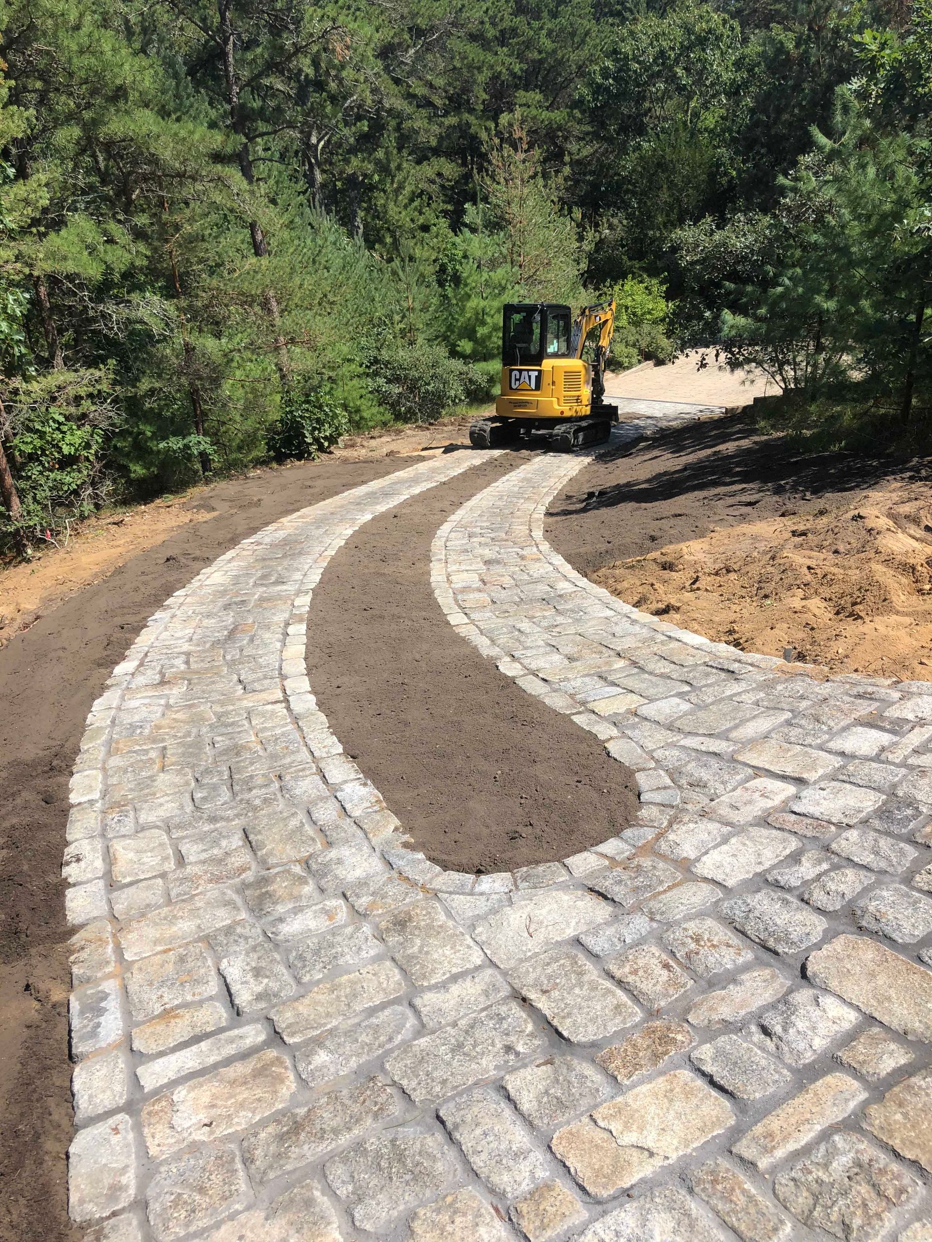 A yellow excavator parked at the end of a winding stone cobblestone driveway surrounded by a wooded area.