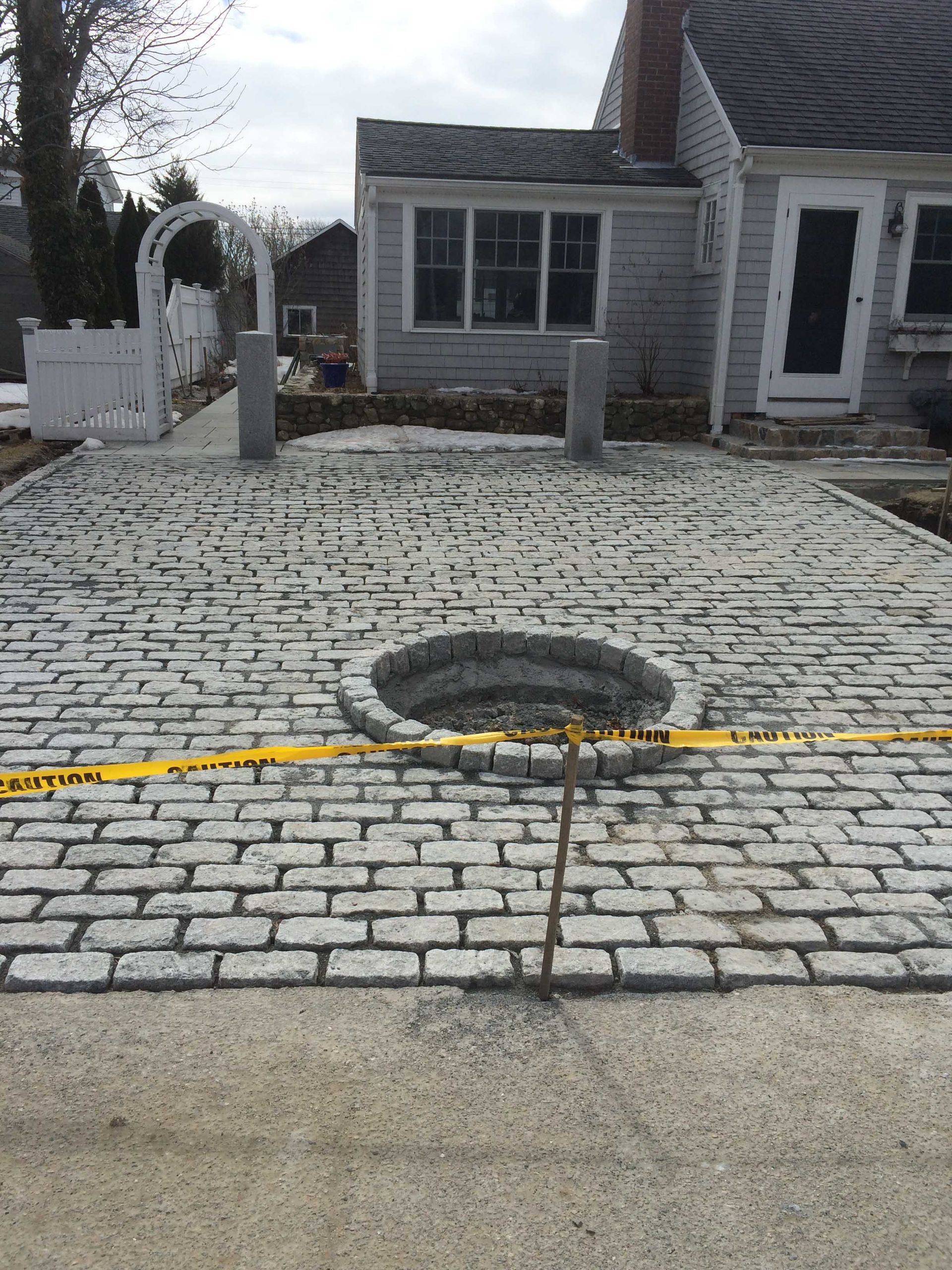 A newly paved gray cobblestone patio featuring a round stone fire pit in the center, fronting a house with yellow tape.