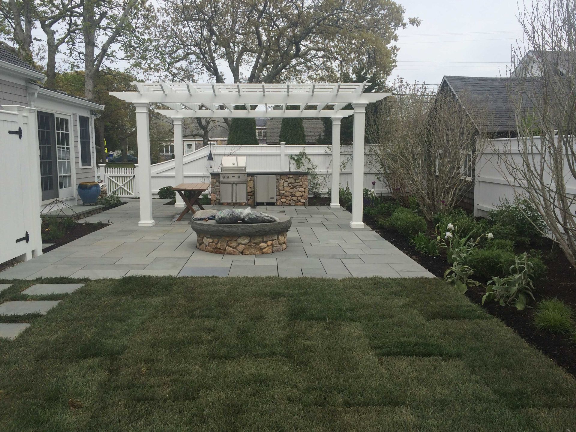 Backyard patio with white pergola, fire pit, paved seating area, and lawn bordered by shrubs.