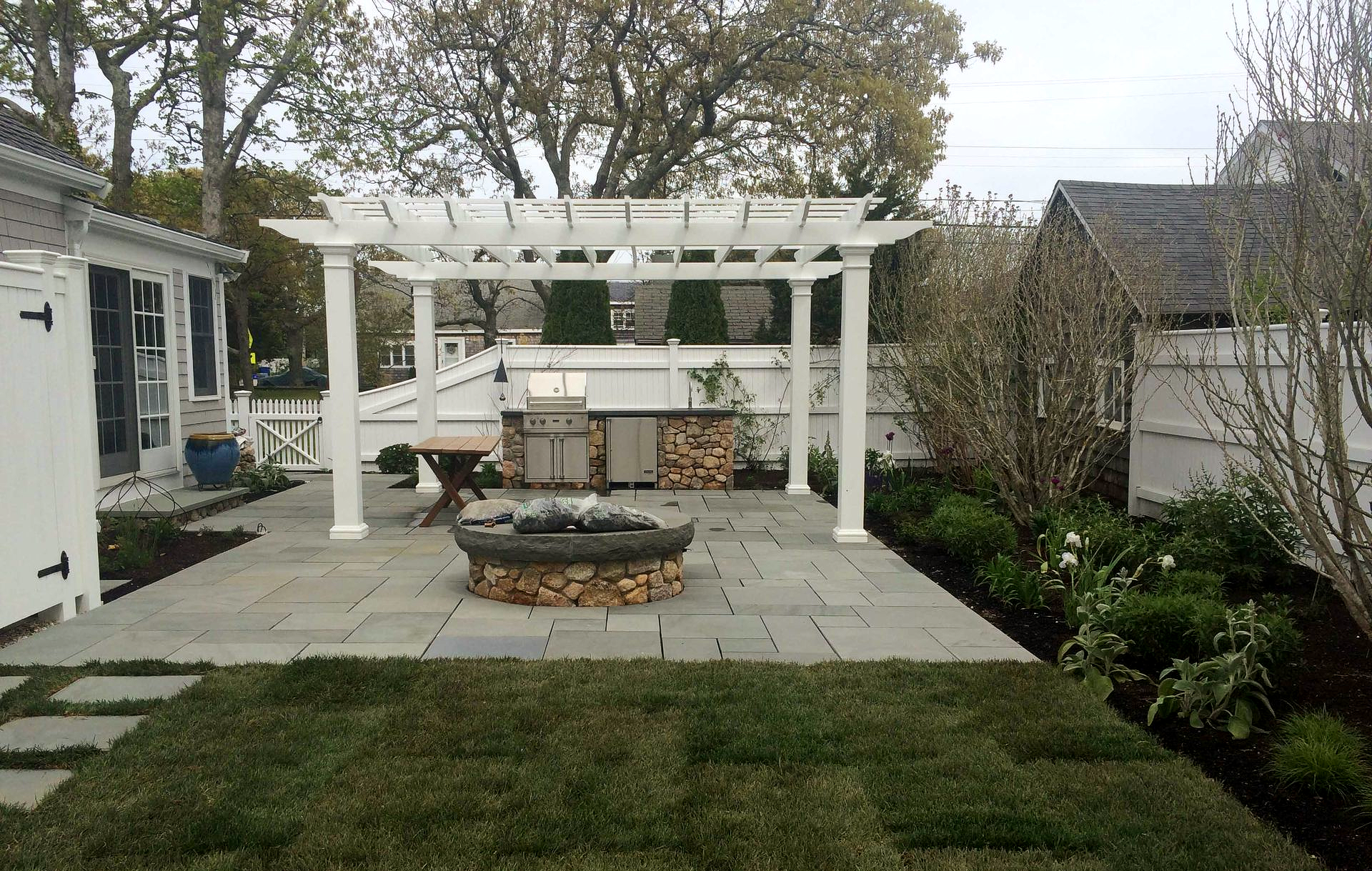 Backyard patio with white pergola, fire pit, stone seating, and landscaped lawn.