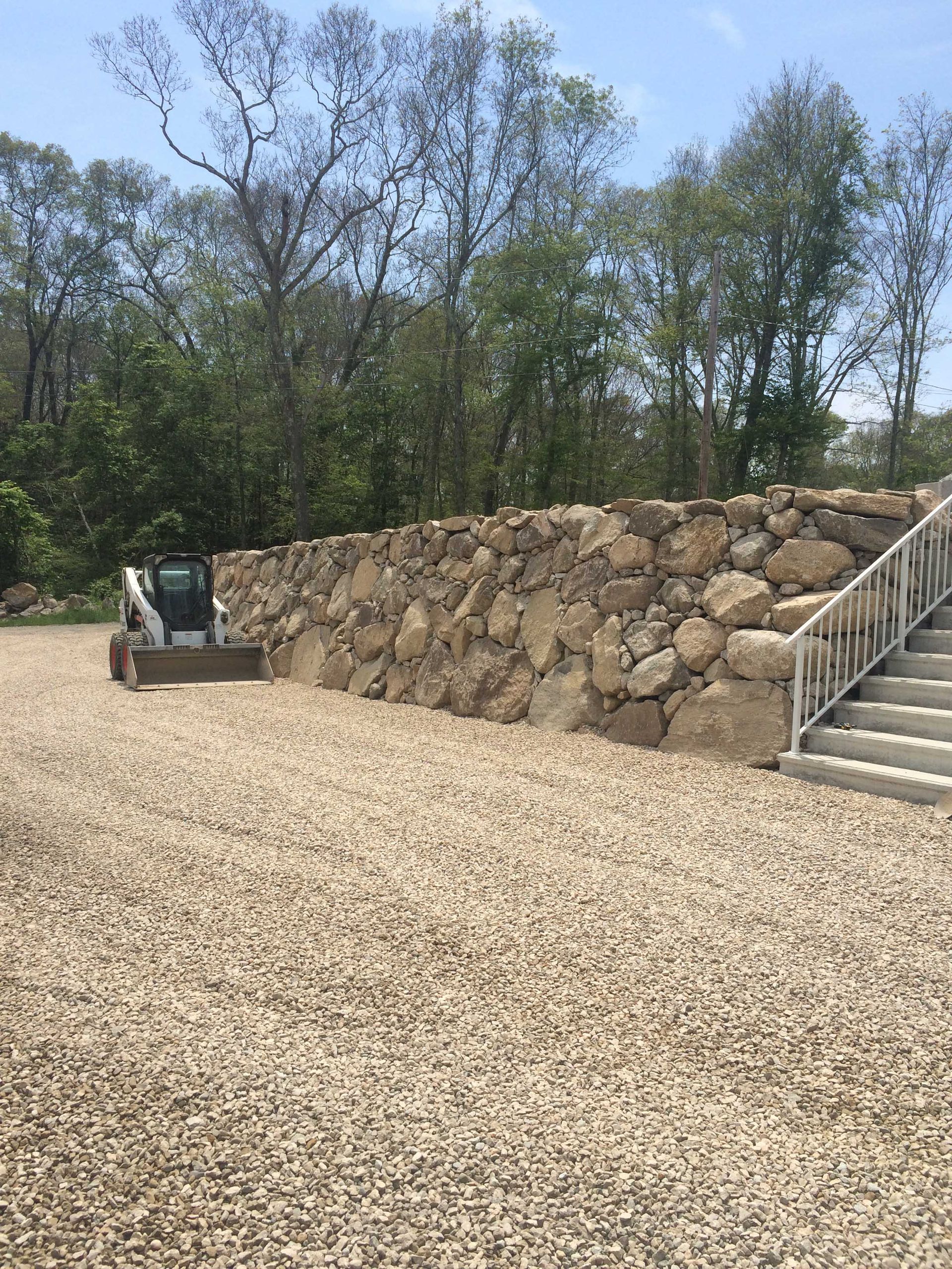 A white skid steer loader parked next to a large natural stone retaining wall near outdoor stairs.