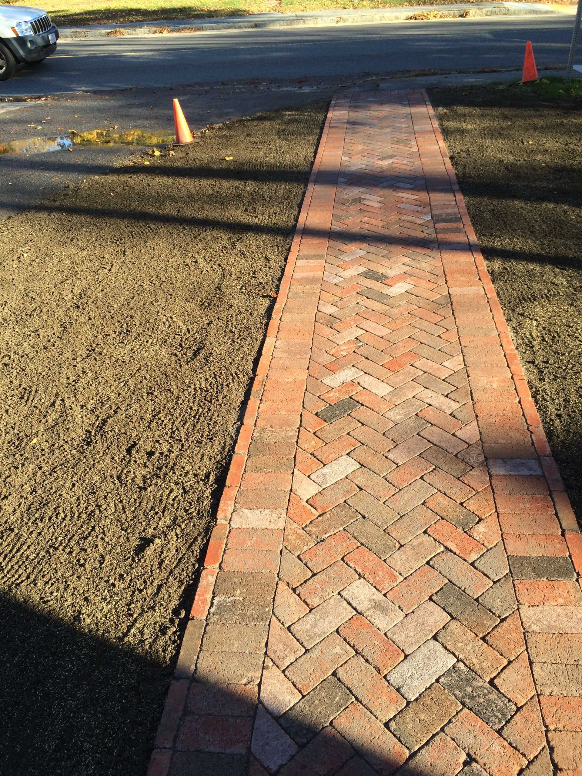 A red brick walkway with a herringbone pattern, flanked by bare soil, leads to a road with two orange traffic cones.