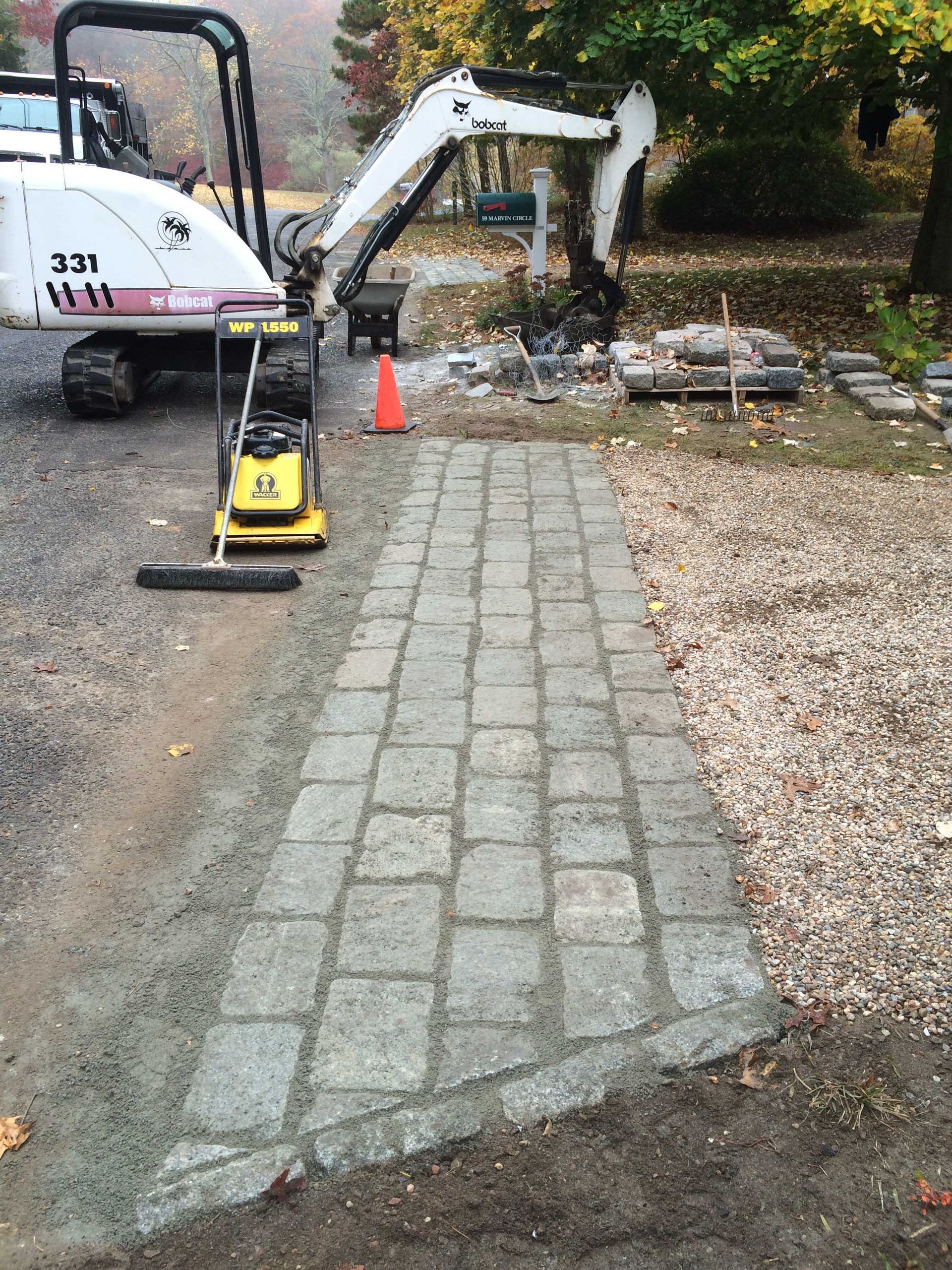 A newly laid stone paver walkway next to a gravel driveway, with a small excavator parked in the background.