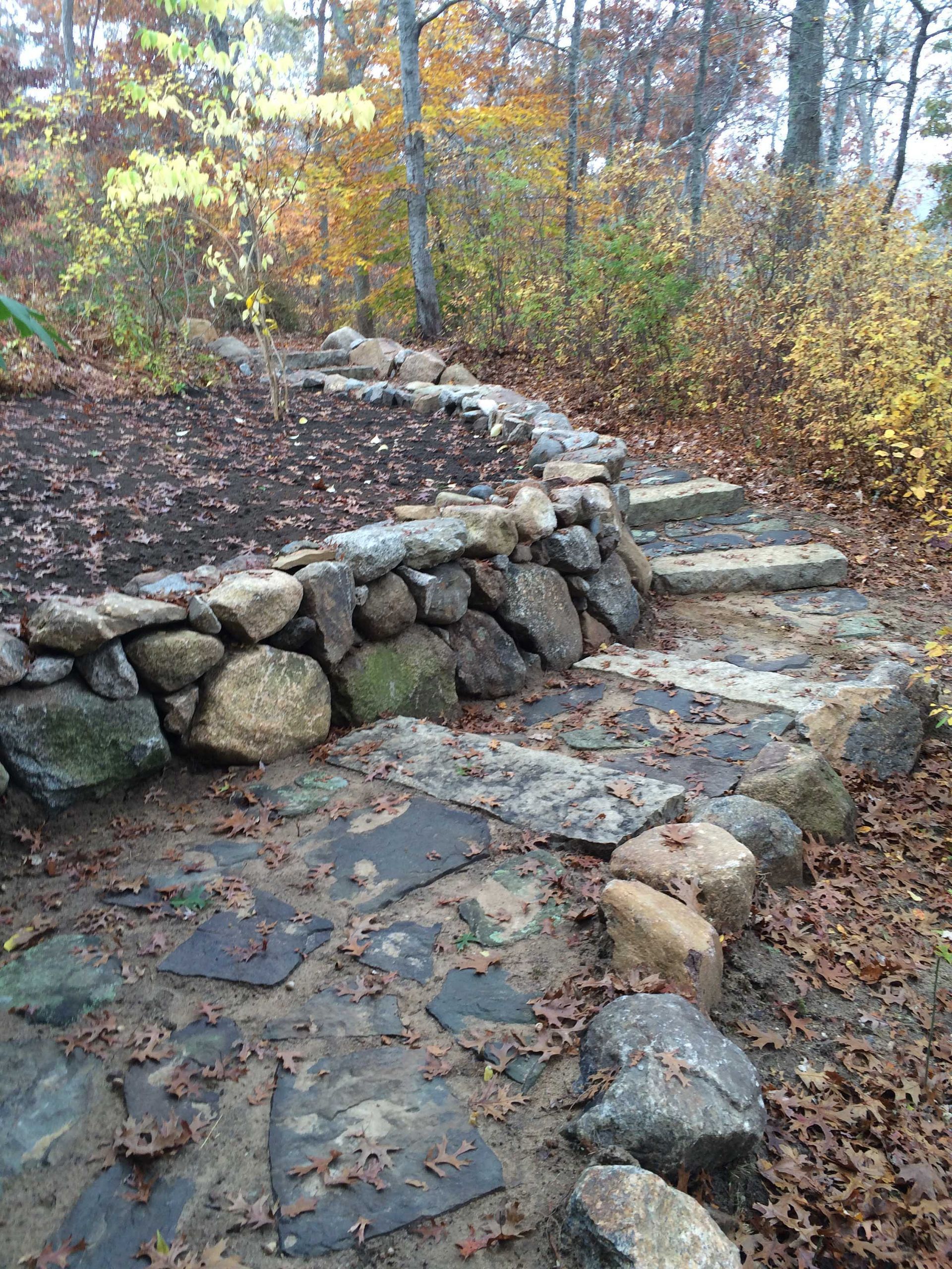 A stone retaining wall curves around a soil garden bed, with stone steps leading up alongside it in a wooded area.