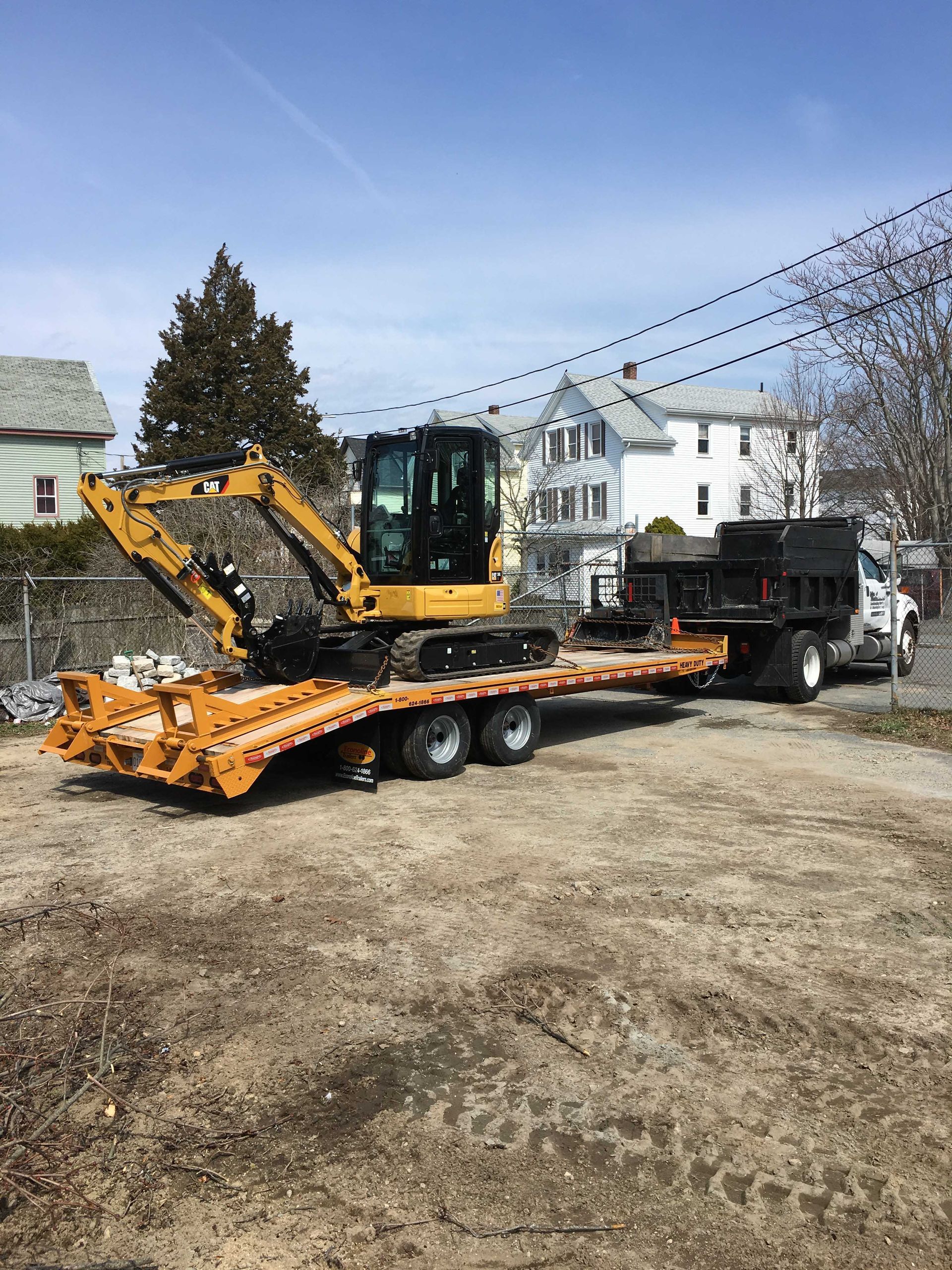 A yellow excavator sits atop a flatbed trailer attached to a dump truck parked on a gravel lot near houses.